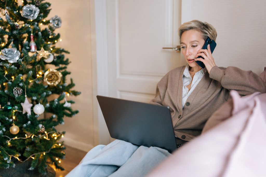 Portrait of busy middle-aged businesswoman remote working on laptop computer, typing on keyboard, looking at screen, talking on mobile phone sitting on couch at Christmas time, xmas tree on background